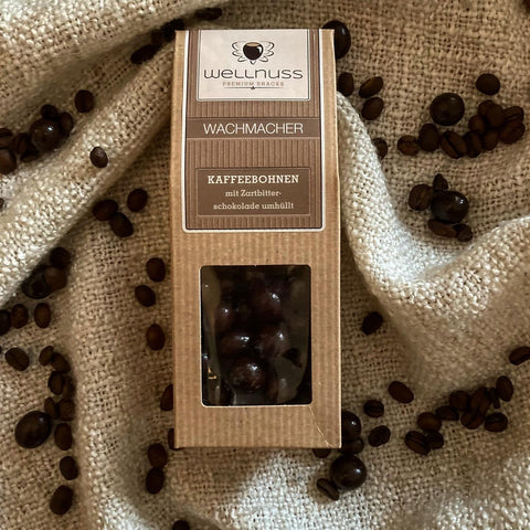 Edle Verpackung der Wachmacher Kaffeebohnen mit Zartbitterschokolade auf beigem Stoff, dekoriert mit Kaffeebohnen. Perfekter Snack für Kaffeeliebhaber.