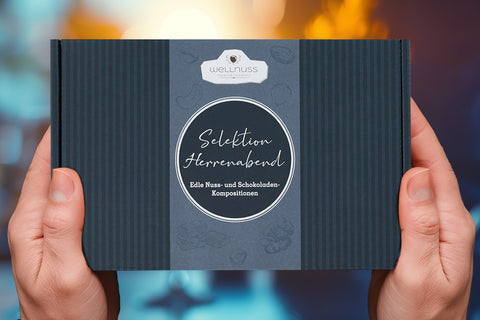 Dieses Bild zeigt eine elegante Geschenkbox mit der Aufschrift „Selektion Herrenabend – Edle Nuss- und Schokoladen-Kompositionen“ von Wellnuss. Die Verpackung hat ein stilvolles Design in dunklen Farbtönen und wird von zwei Händen gehalten. 