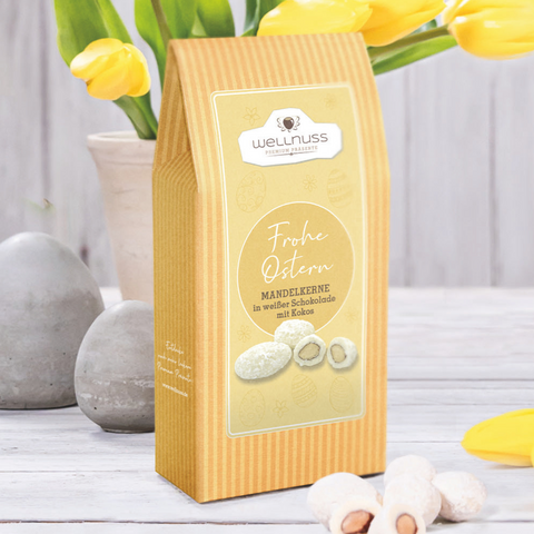 Maxi Faltschachtel Oster Kokosmandeln in weißer Schokolade, 175g, dekorativ präsentiert mit gelben Tulpen und Ostereiern.