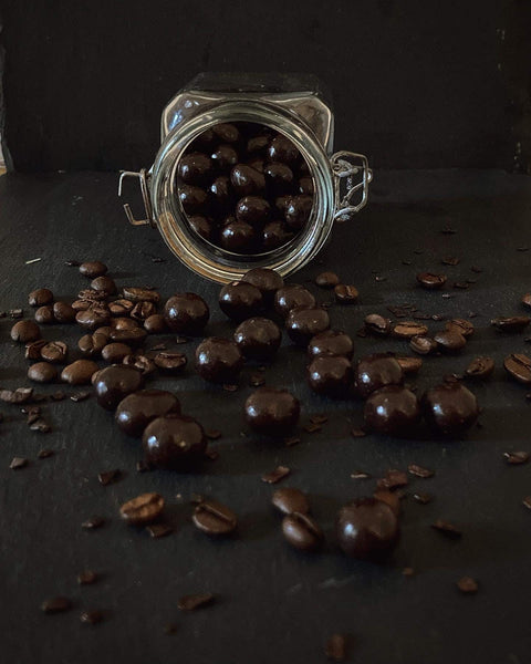 Offenes Glas mit Wachmacher Kaffeebohnen in Zartbitterschokolade, umgeben von Kaffeebohnen auf dunklem Untergrund. Genussvolle Kaffeekombination.